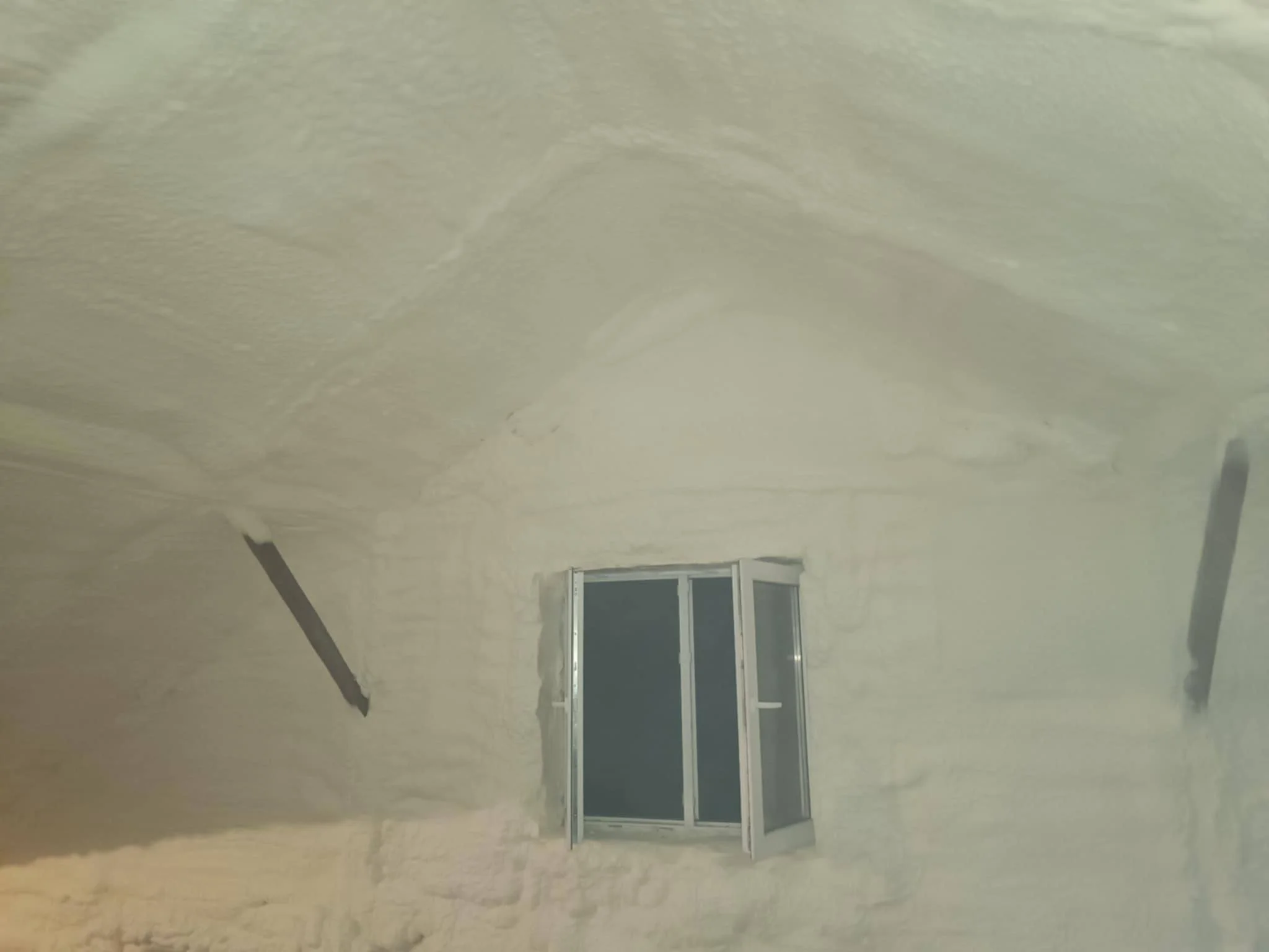 A room with smooth, white spray foam insulation on the walls and ceiling, a small double-pane window, and black support beams.