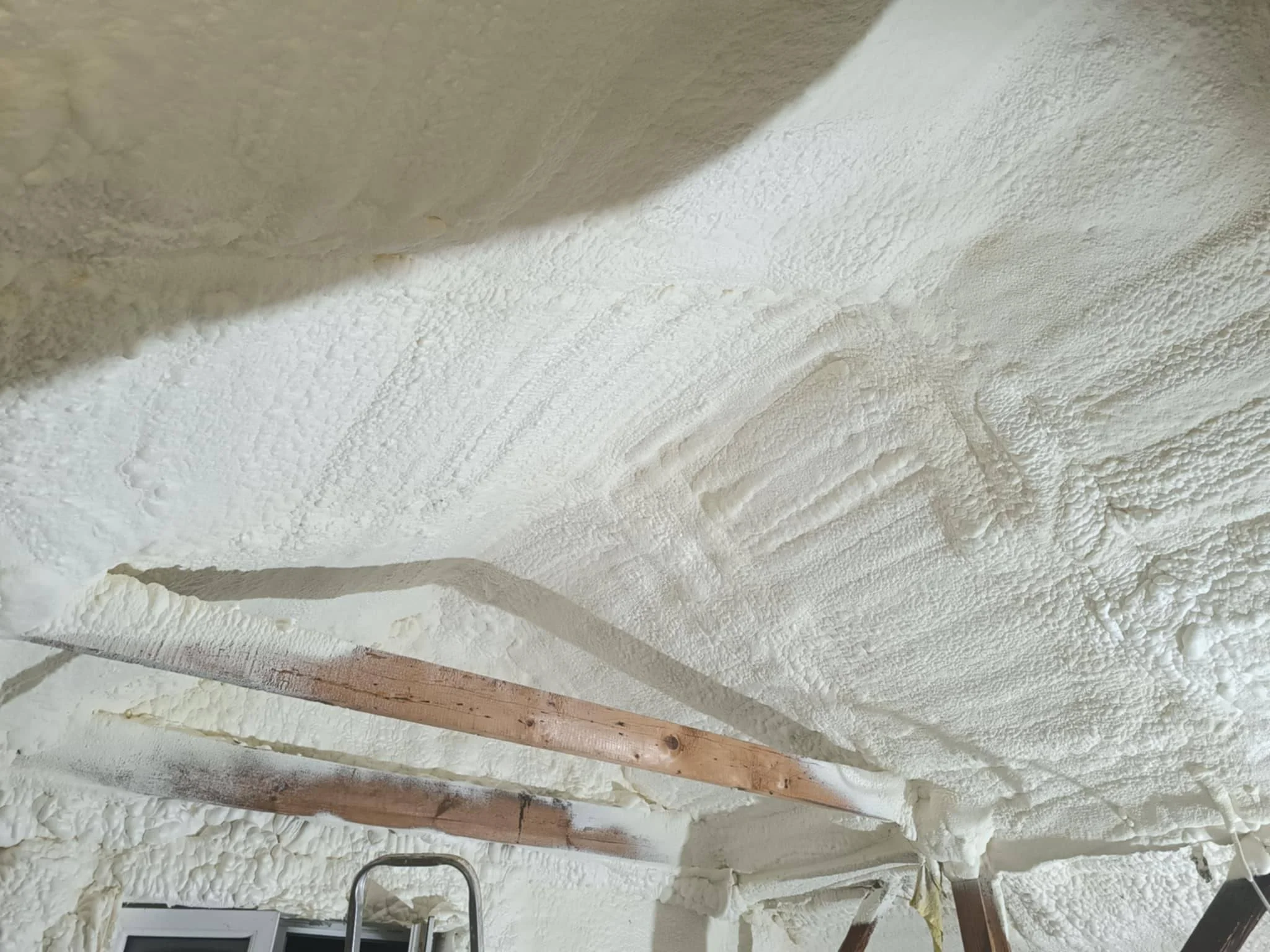 Ceiling covered in spray foam insulation with wooden beams and a ladder visible at the bottom.