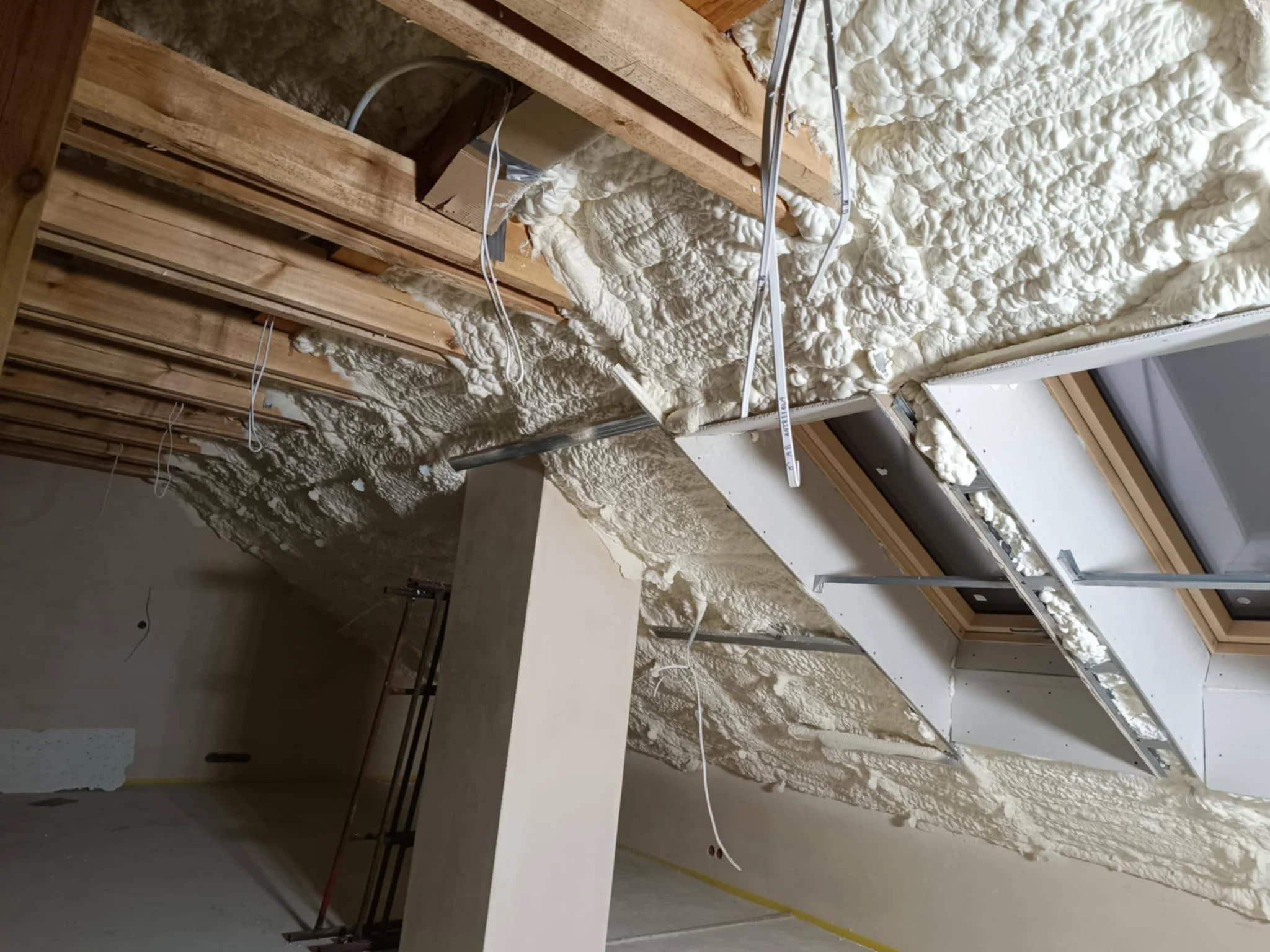 Construction site showing a wall with drywall and spray foam insulation, electrical wiring, and a window, during renovation or building process.