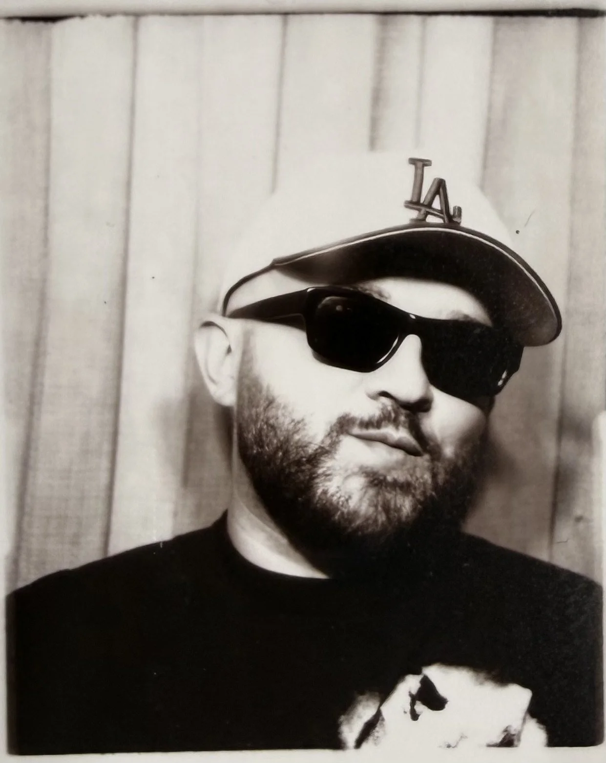 Black and white portrait of a man with a beard wearing a baseball cap with "LA" logo and sunglasses, standing in front of a wooden wall.