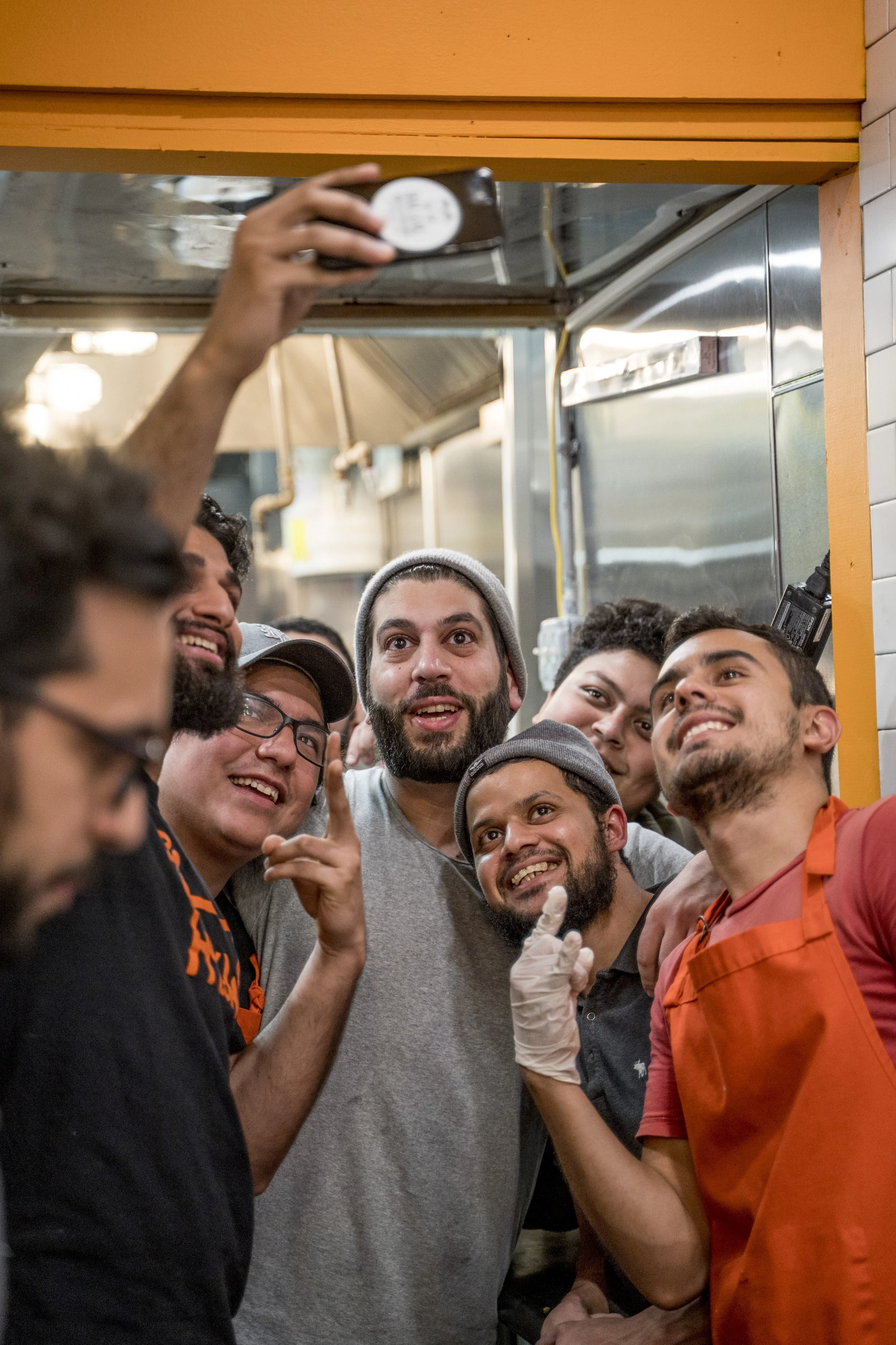A group of people in a kitchen, taking a selfie together. Some are smiling and looking at the camera, one person is wearing an orange apron, and another is wearing gloves.