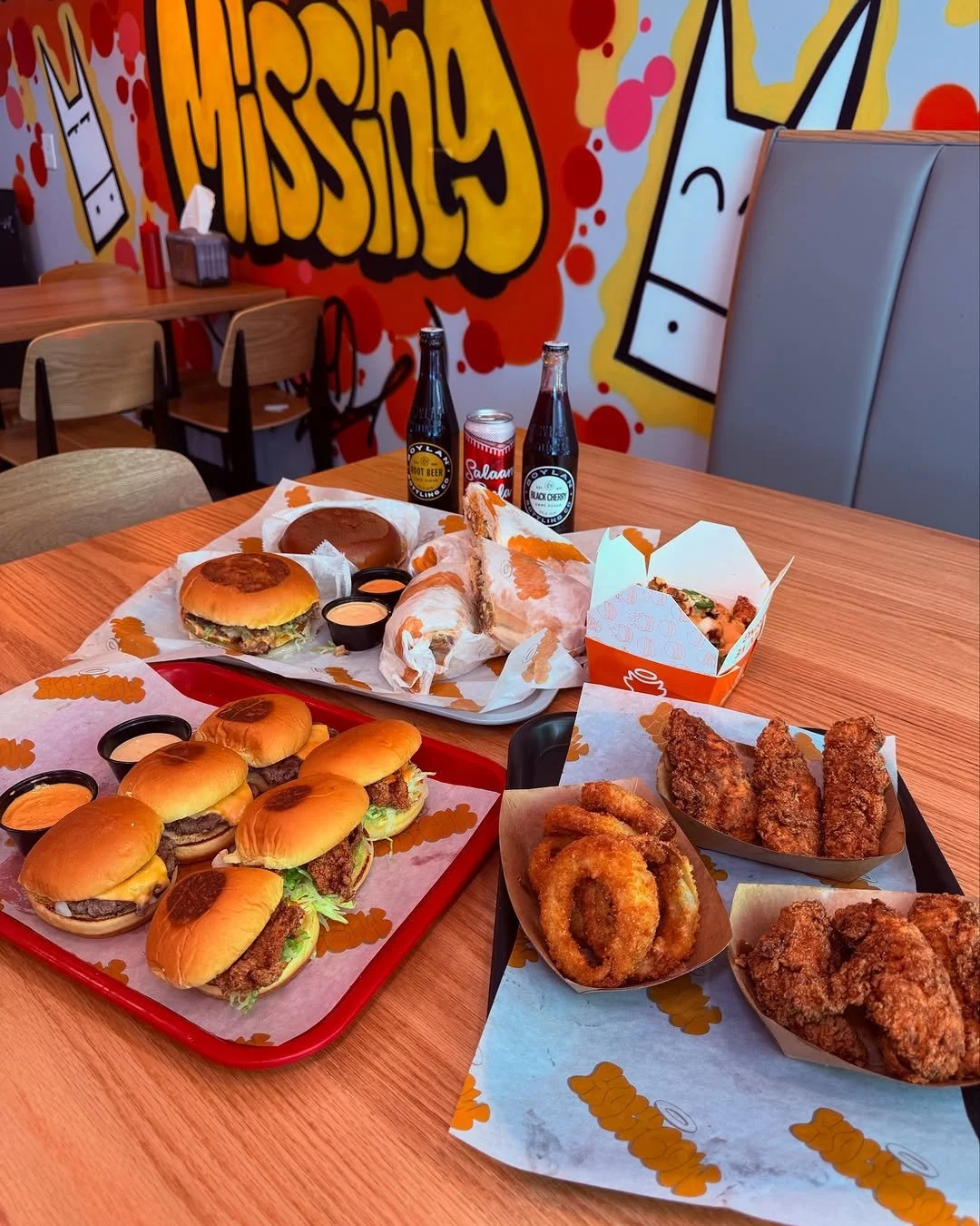 A table filled with fast food including burgers, fried chicken pieces, onion rings, and drinks, in a restaurant with a colorful graffiti wall.