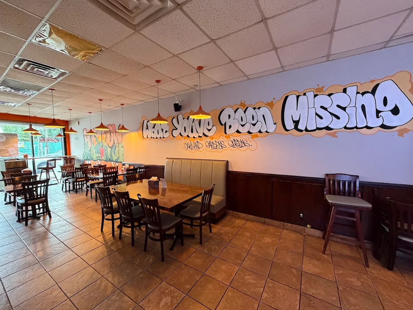 Interior of a restaurant with several wooden tables and chairs, a colorful mural on the wall that says 'LOL & WOW' with large leafy green plants and a red heart, hanging orange pendant lights, and a large window letting in natural light.