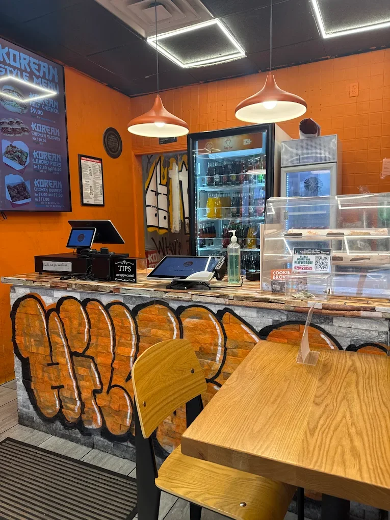 Graffiti-style art on restaurant wall featuring the phrase 'Sam That Food' in orange and black, with modern pendant lights hanging from the ceiling, wooden tables with chairs, and a large glass window showing outdoor parking.