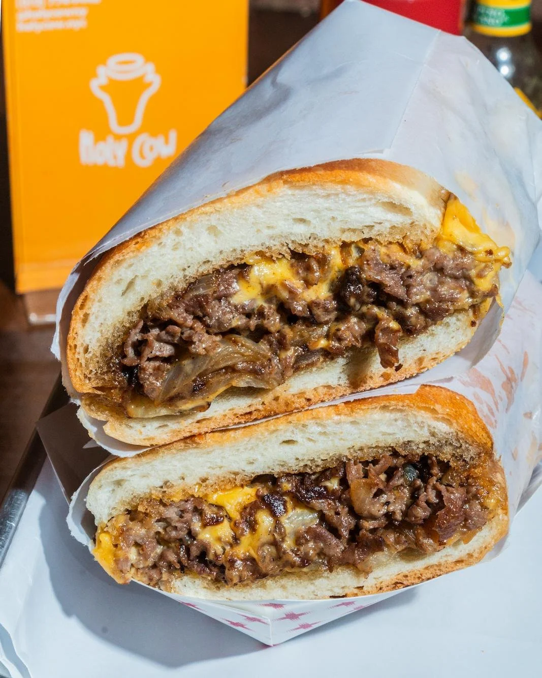A Beef and Cheddar sandwich cut in half, showing chopped beef, melted cheese, and caramelized onions inside a soft bun, wrapped in white paper.