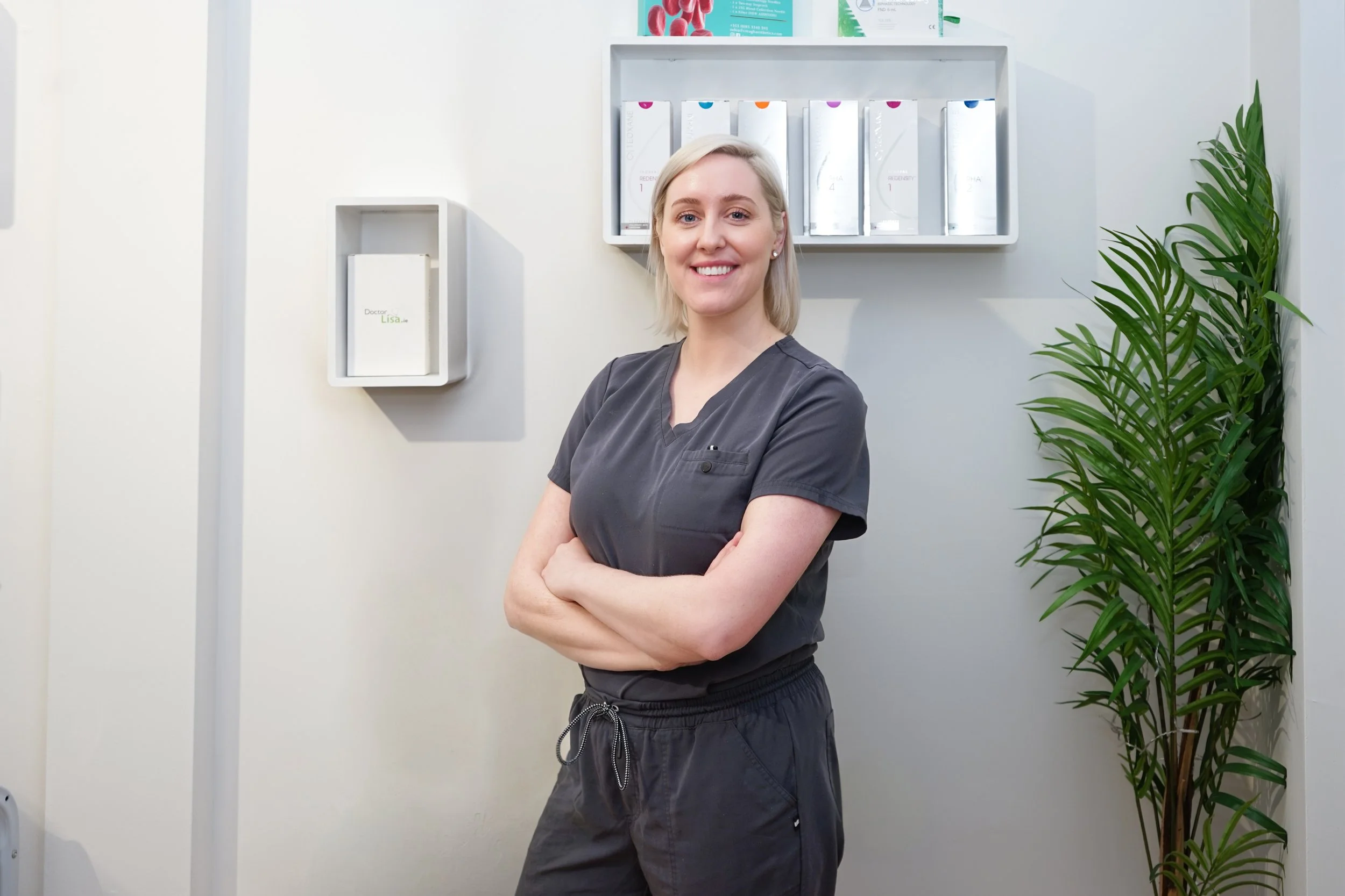 A smiling female healthcare professional with blonde hair, wearing a dark gray medical scrub, standing with arms crossed in a medical office or clinic. Behind her are framed certificates or medical information on the wall, and a tall green plant on her right.