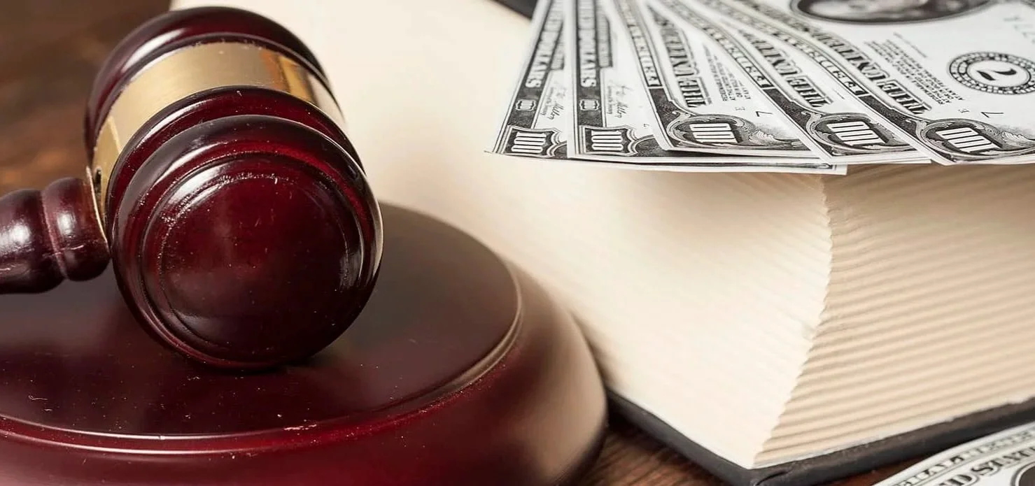 Gavel resting on a round sounding block, with a thick book and several hundred-dollar bills on top of the book.