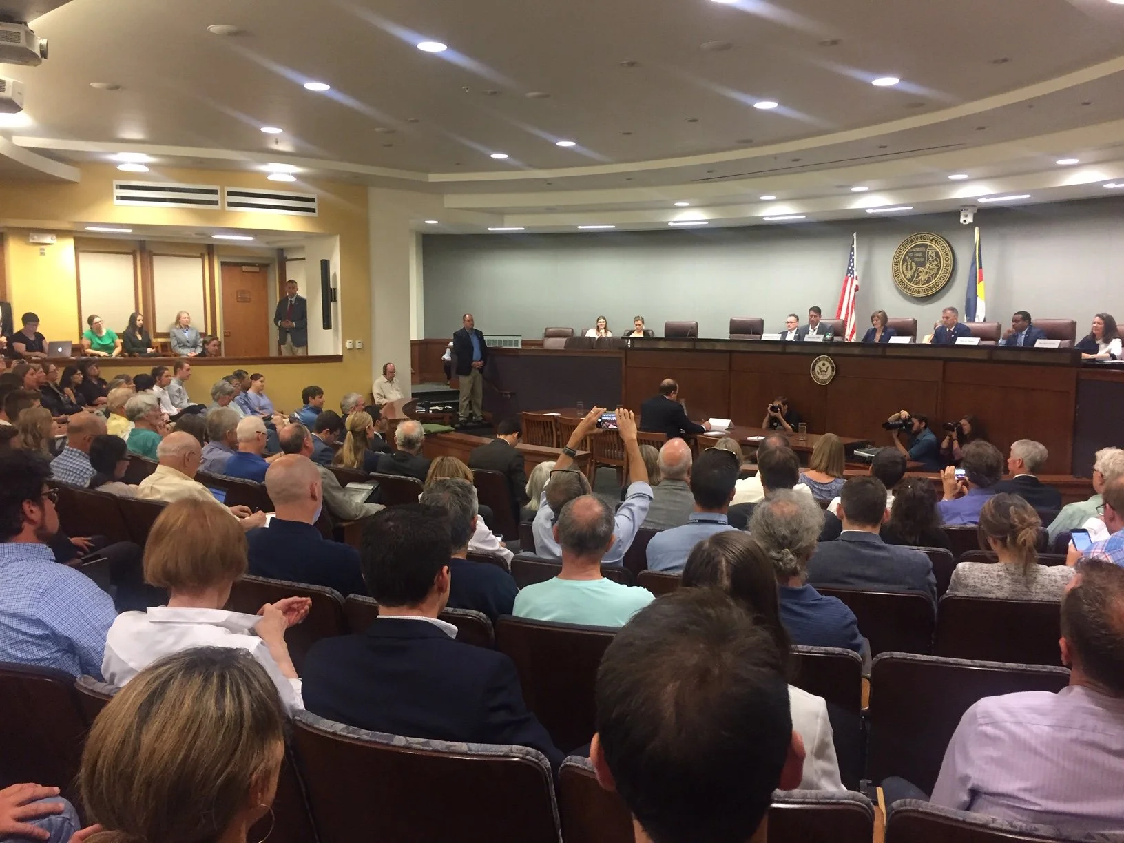 A crowded courtroom filled with people seated and listening to a panel of officials seated at a long raised desk. The room has flags, a seal, and a mix of seated attendees and reporters taking photos and notes.