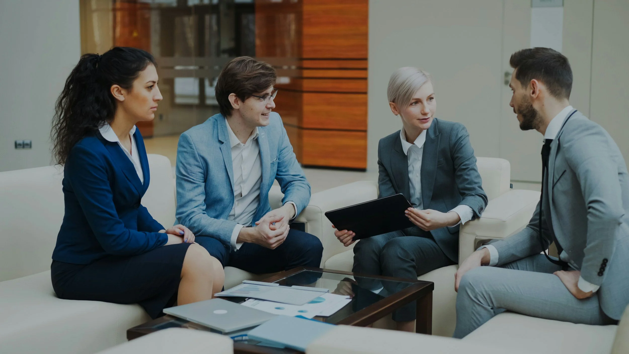 Five professionals in business attire having a discussion in a modern office lounge.