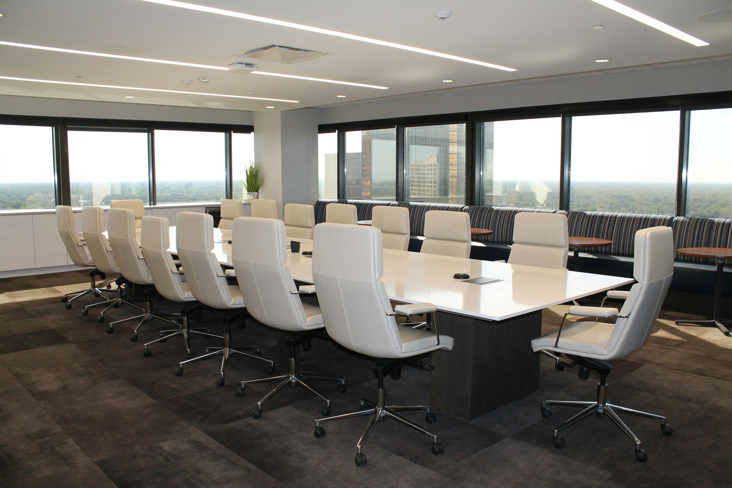 Modern conference room with a long white table surrounded by white office chairs, large windows with city and skyline view, and a seating area with striped cushions along the windows.