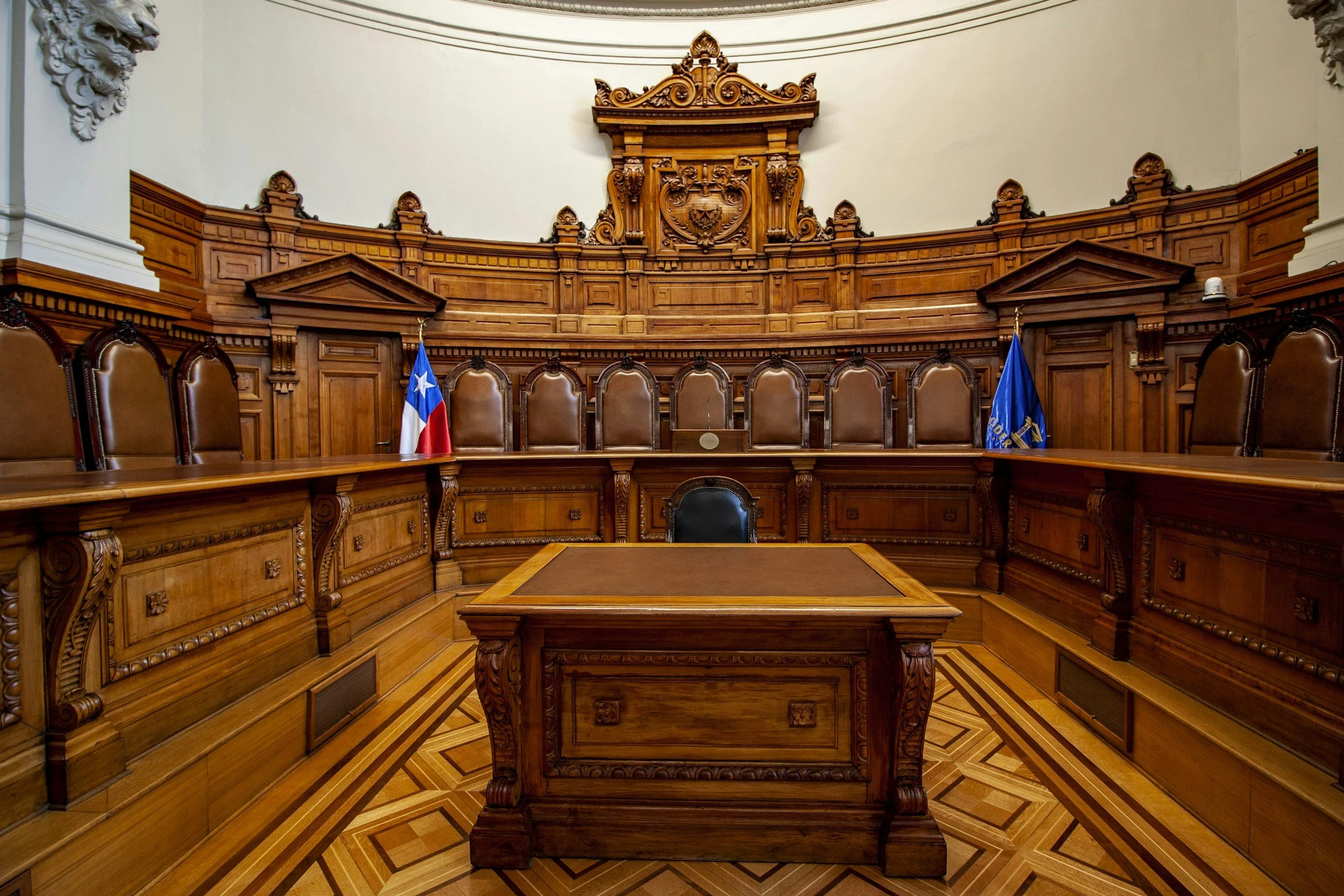 A historic courtroom with wooden paneling and high-backed leather chairs, featuring the Chilean flag on the left and a blue flag on the right.