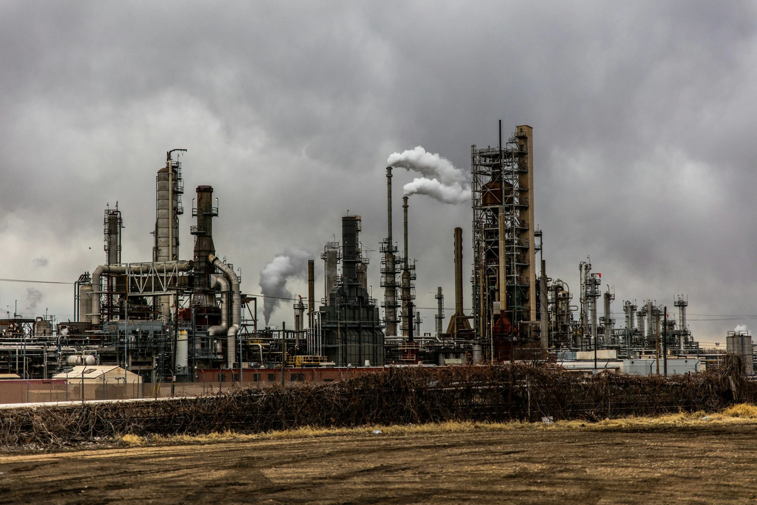 An industrial oil refinery with numerous pipes, towers, and smokestacks emitting smoke under a dark, cloudy sky.