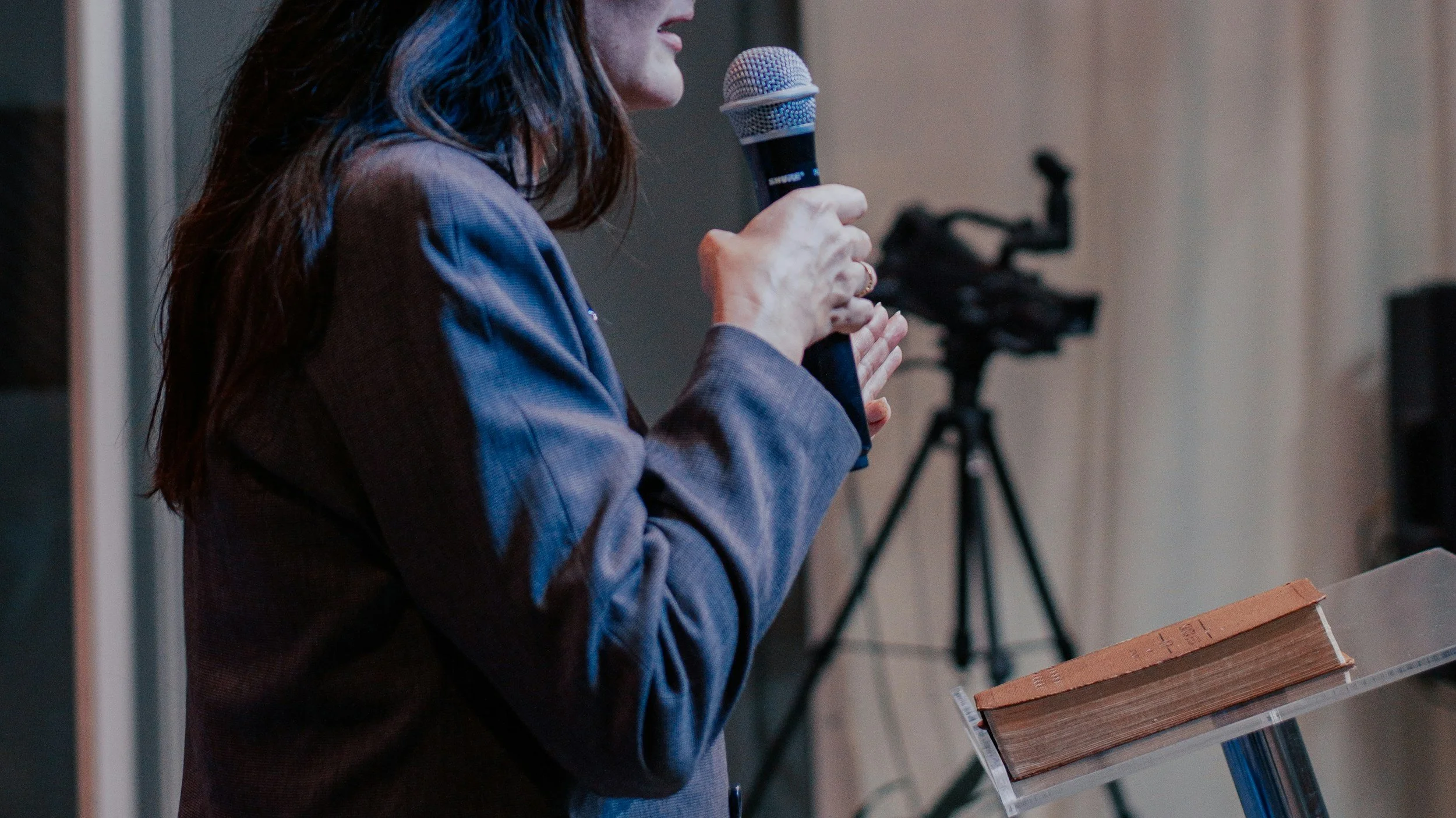 A woman in a blazer speaking into a microphone at a lectern with an open book, with a camera on a tripod in the background.