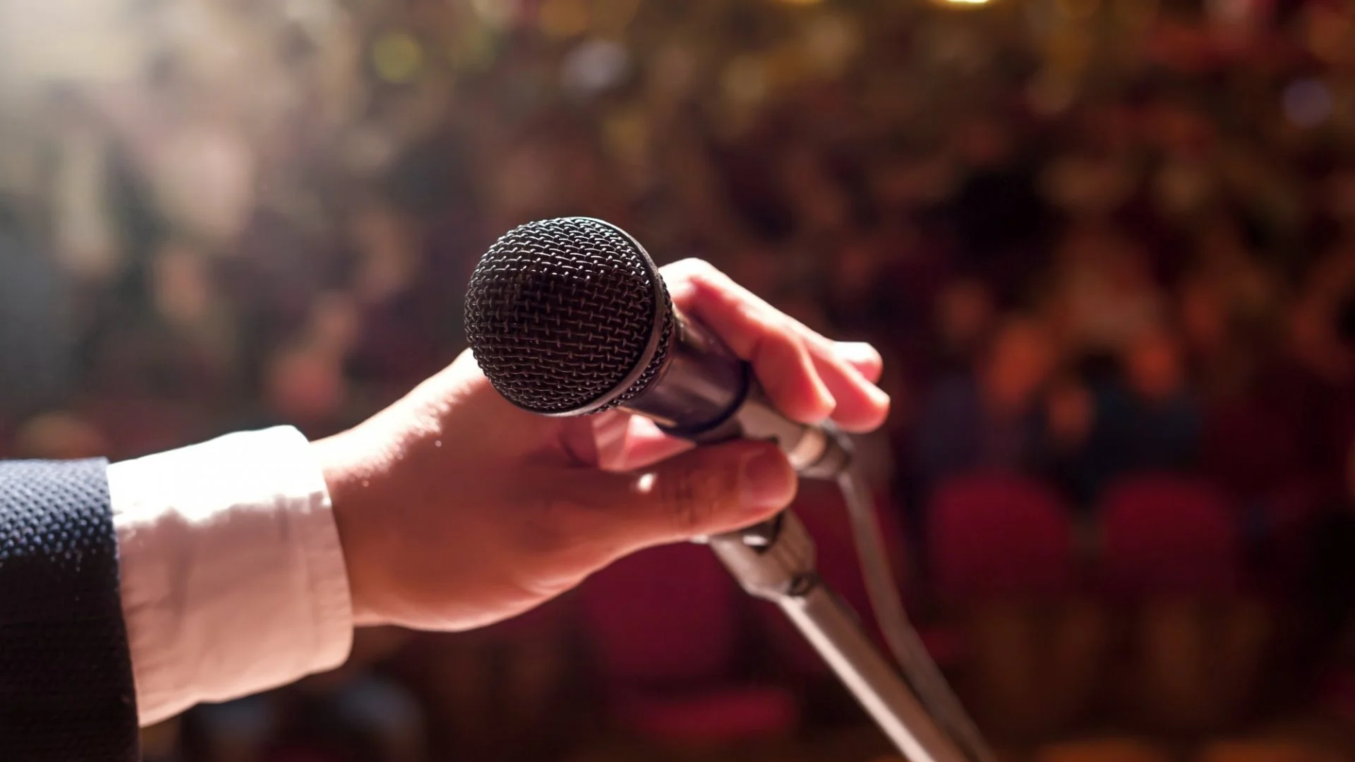 A person's hand holding a microphone, with a blurred background of audience or audience seats.