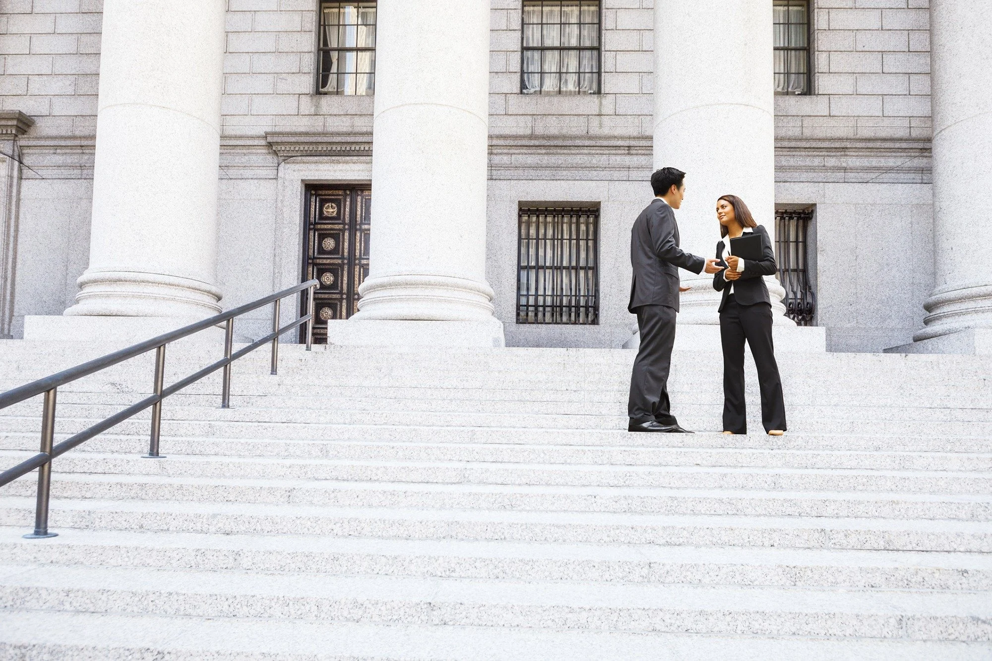 Two businesspeople, a man and a woman, dressed in black suits, standing and talking on the steps of a large neoclassical building with tall white columns.