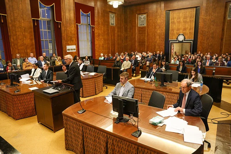 A courtroom filled with people; a man standing at a podium appears to be speaking or presenting, with several officials seated at a table with monitors and paperwork, and a large audience seated behind.