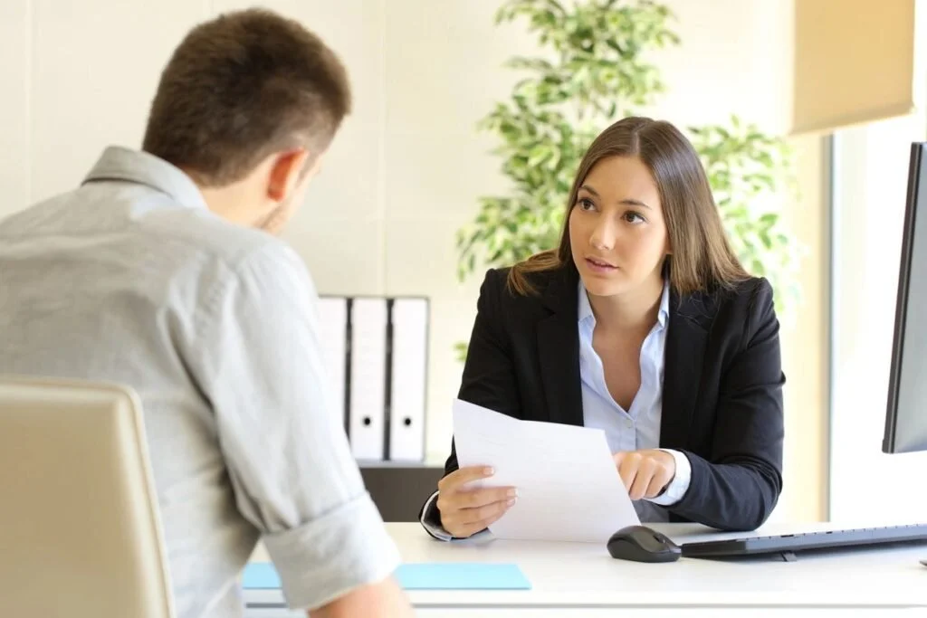 A woman in a business suit sitting at her desk, talking to a man in a light-colored shirt, holding documents in an office setting.