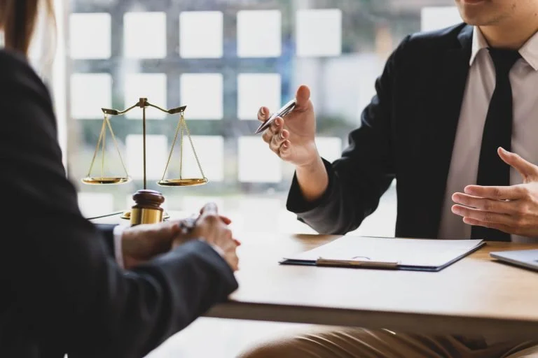 Two people in business attire having a discussion at a table, with a legal scale and gavel in the background.