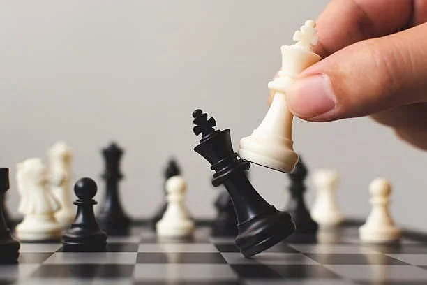 Close-up of a hand moving a black king chess piece on a chessboard with other white and black pieces in the background.