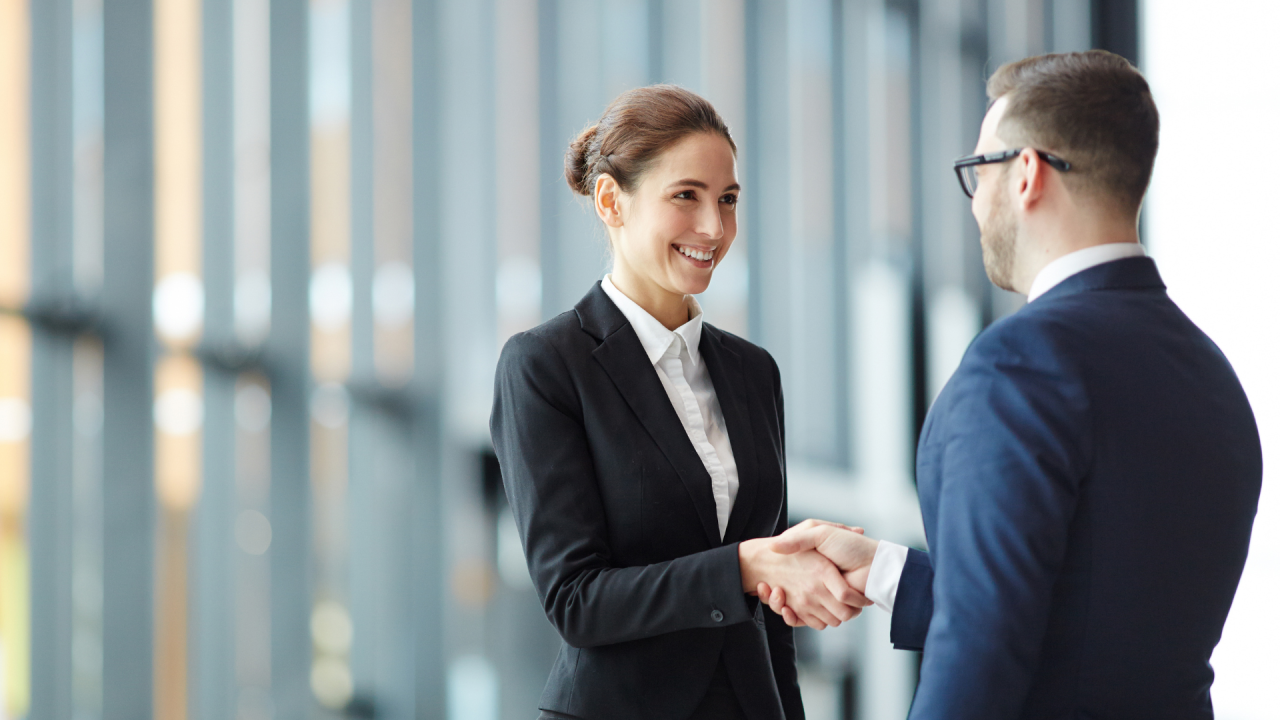 A woman and a man shaking hands in a professional setting with large windows in the background.