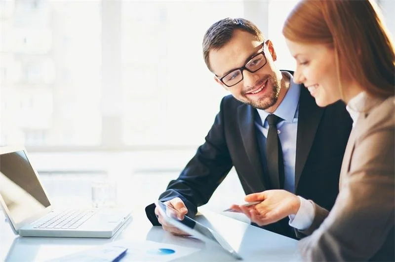 Two colleagues, a man with glasses and a woman, smiling and looking at a tablet in a bright office.