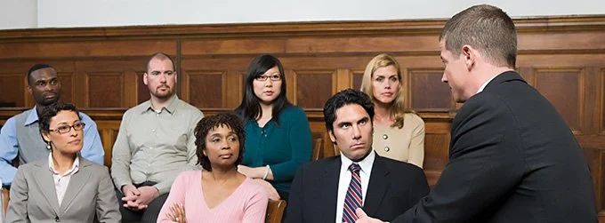 A man in a suit is speaking to a group of diverse people sitting in a courtroom or formal setting, with wooden paneling in the background.