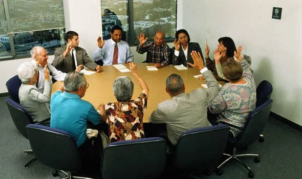 A diverse group of eleven people in a business meeting around a large oval table in a conference room. Some participants are raising their hands, possibly voting or contributing to a discussion. There are papers and pens on the table, and a large window in the background shows a cityscape.