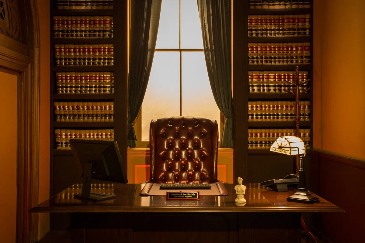A vintage office with a large leather boss chair behind a dark wood desk, a computer monitor to the left, a lamp and a small bust statue on the right, and a nameplate reading 'Surgeon John Smith' in the center, with tall bookshelves filled with legal books flanking a window with curtains in the background.