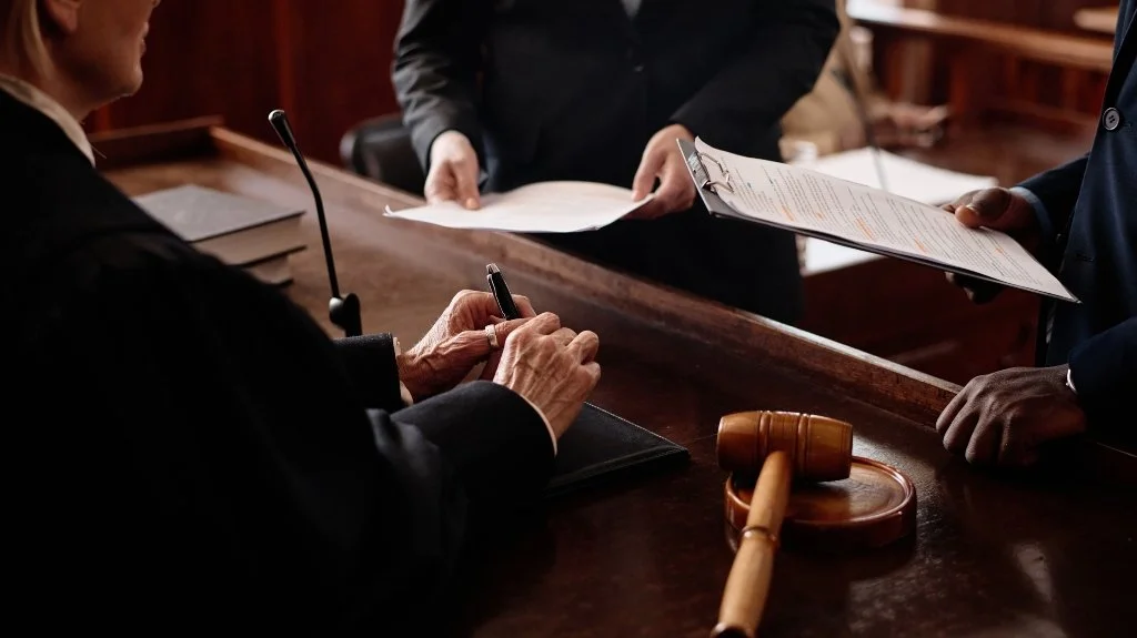 Legal proceeding with a judge's gavel, a woman attorney signing documents, and other attorneys holding papers in a courtroom.