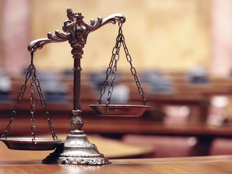 A decorative brass balance scale on a wooden surface, with a blurred courtroom background.