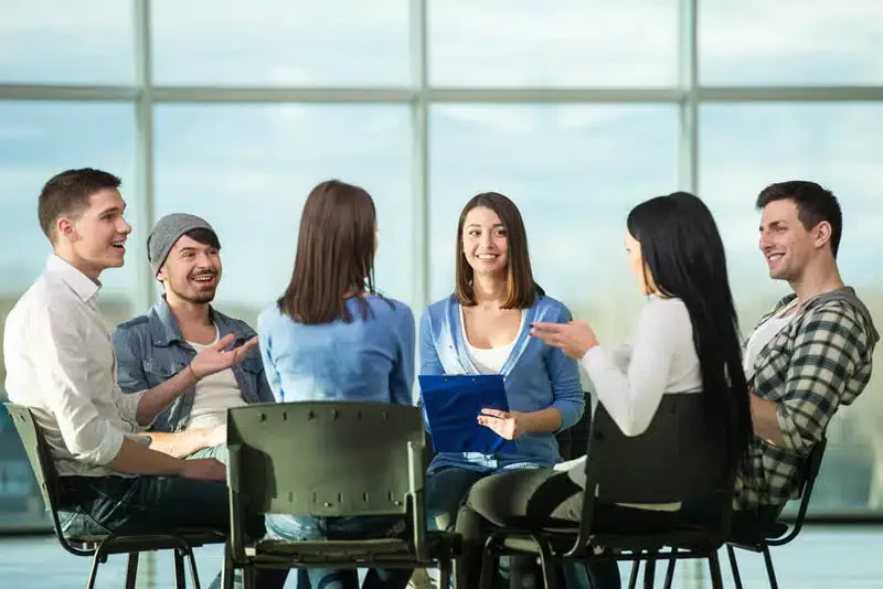 Group of six young adults gathered around a table in a modern, glass-walled room, engaged in conversation and smiling.