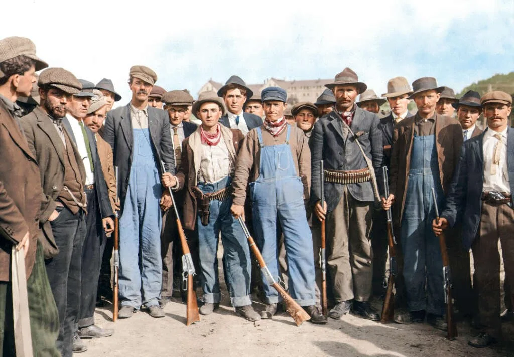A large group of men dressed in early 20th-century rural or industrial work attire, some holding rifles, standing outdoors with a clear sky and some buildings in the background.