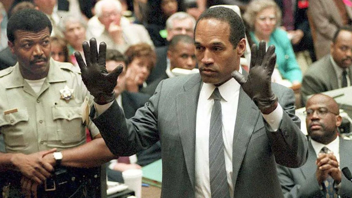 A man in a gray suit and tie with black gloves raising both hands during a court proceeding, with several people seated behind him, including a police officer.