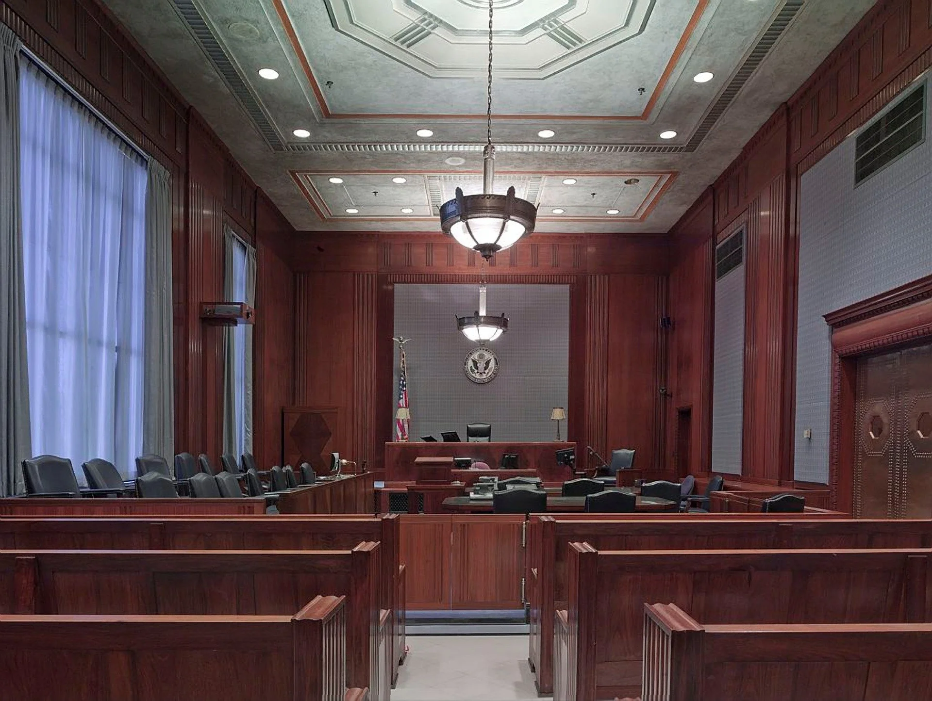 View of a courtroom with wooden paneling, a judge's bench, empty chairs, the American flag, and the United States seal on the wall behind the bench.
