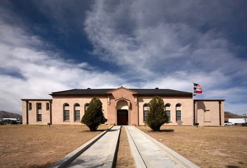 A large, historic-style building with a pink stucco exterior, arched windows, and a central entrance. Two trees are in front, and a paved walkway leads to the entrance. An American flag is flying on a flagpole to the right. The sky is partly cloudy with dark clouds and blue sky visible.