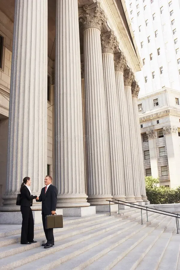 Two business people, a woman and a man, shaking hands on the steps in front of a building with large, ornate Greek-style columns.