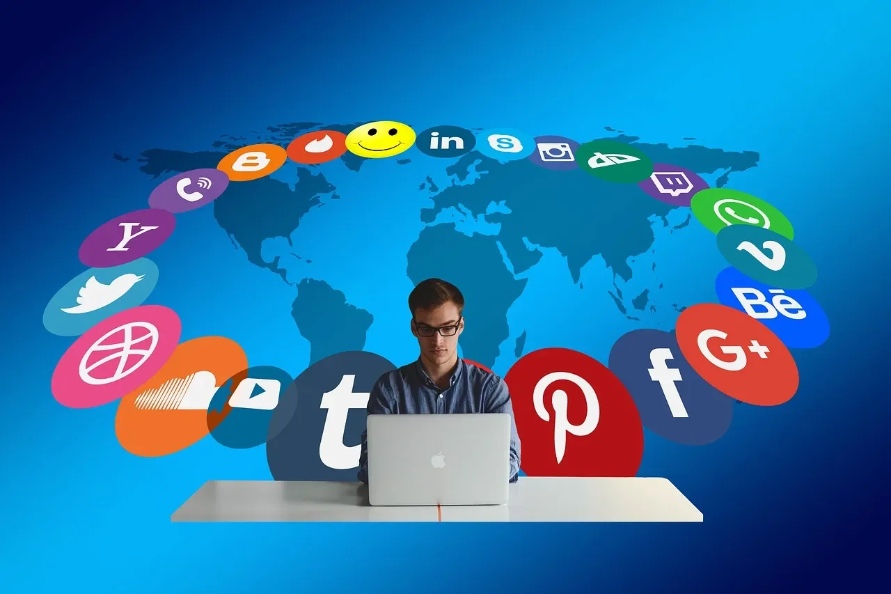 A young man working on a laptop at a desk with a background of a world map and floating social media icons such as Facebook, Twitter, Instagram, LinkedIn, WhatsApp, and YouTube.