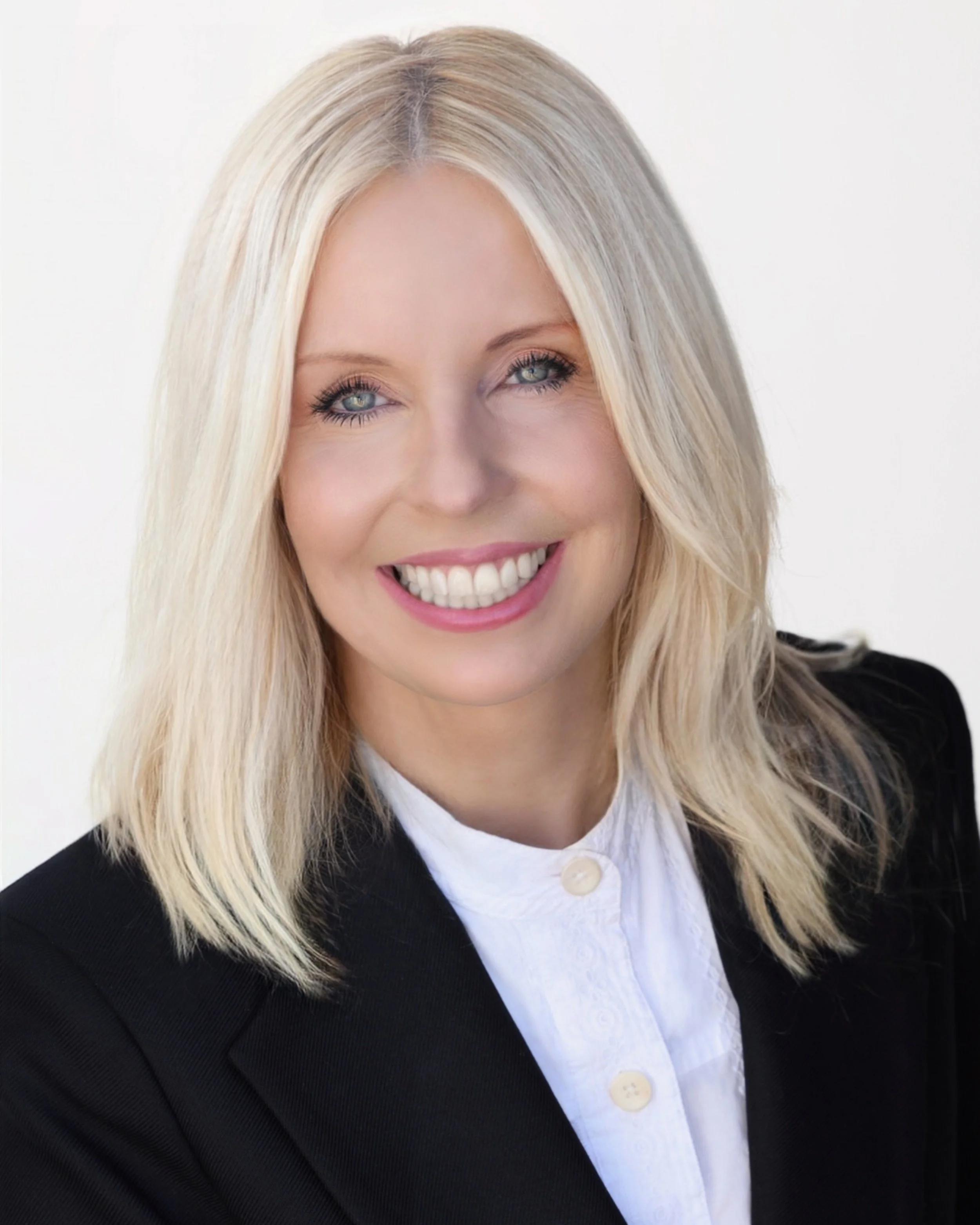 A woman with blonde hair and blue eyes smiling, wearing a black blazer and white blouse, on a white background.