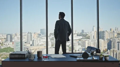 A man in a suit standing at a large office window overlooking a cityscape.