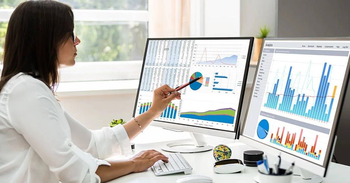 Woman working at desk with dual computer monitors displaying charts and graphs, pointing at one monitor with a pen.