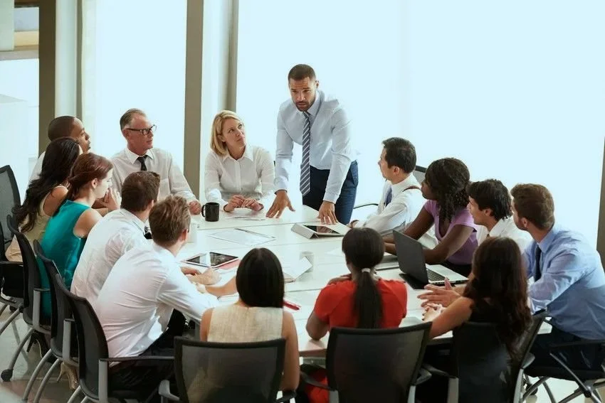A business meeting with a diverse group of people gathered around a conference table, with one man standing and speaking to the group.