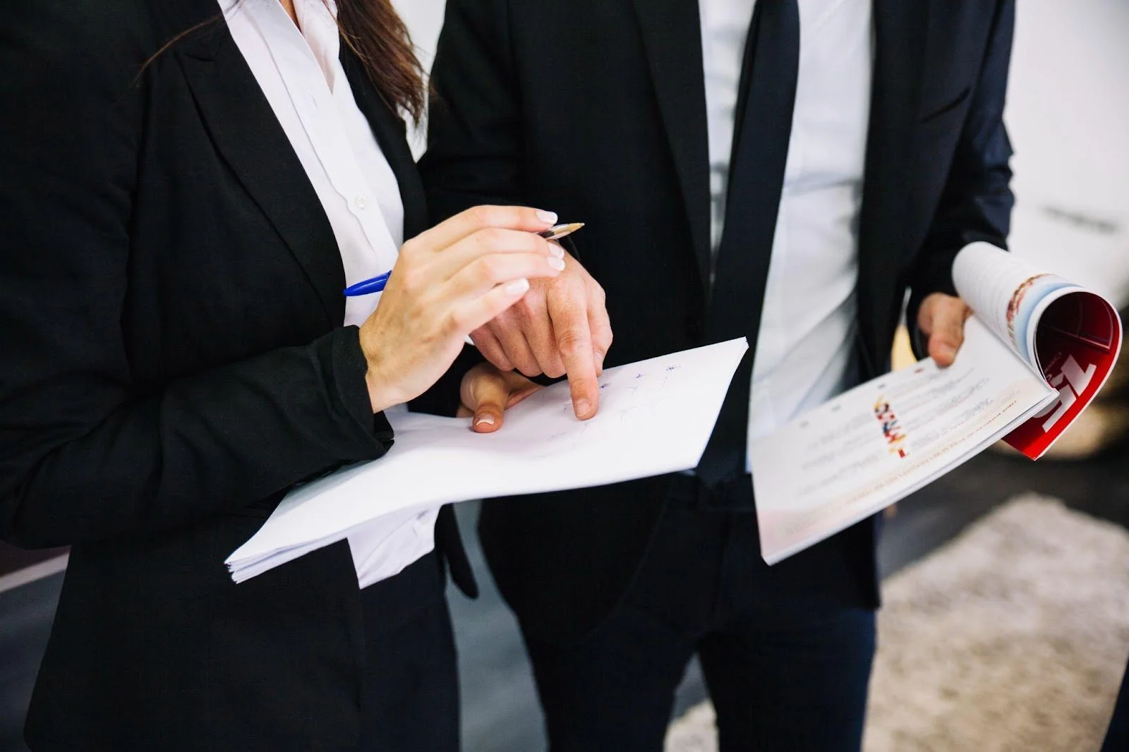 Two people in formal business attire examining documents, with one pointing at a paper.