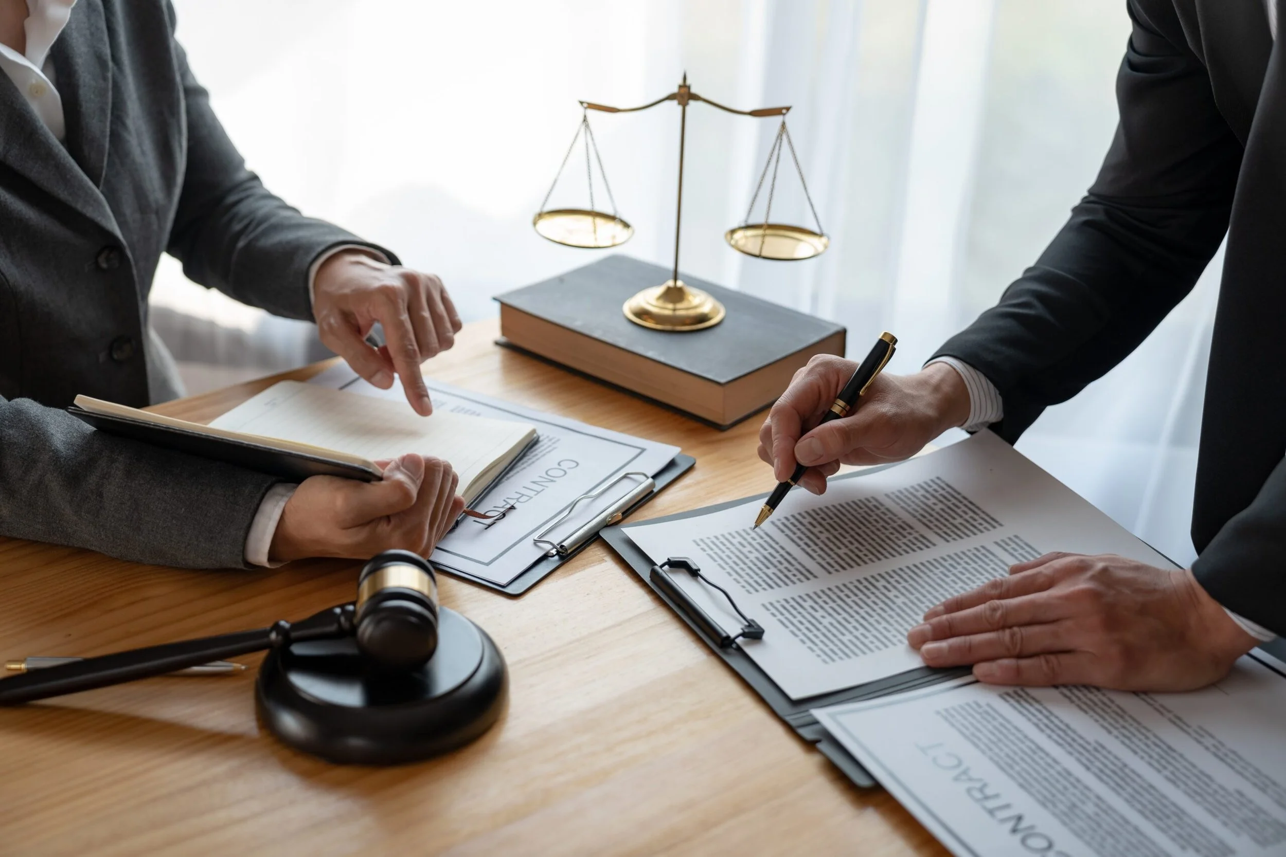 Two people in formal attire reviewing documents on a wooden table, with legal symbols like a gavel, a scale of justice, and law books.