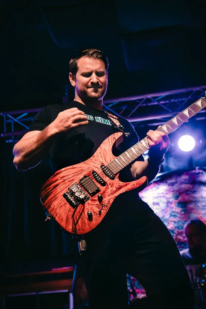 A man playing an electric guitar on stage at Fat Fish Blue, with musical equipment and a sign in the background.