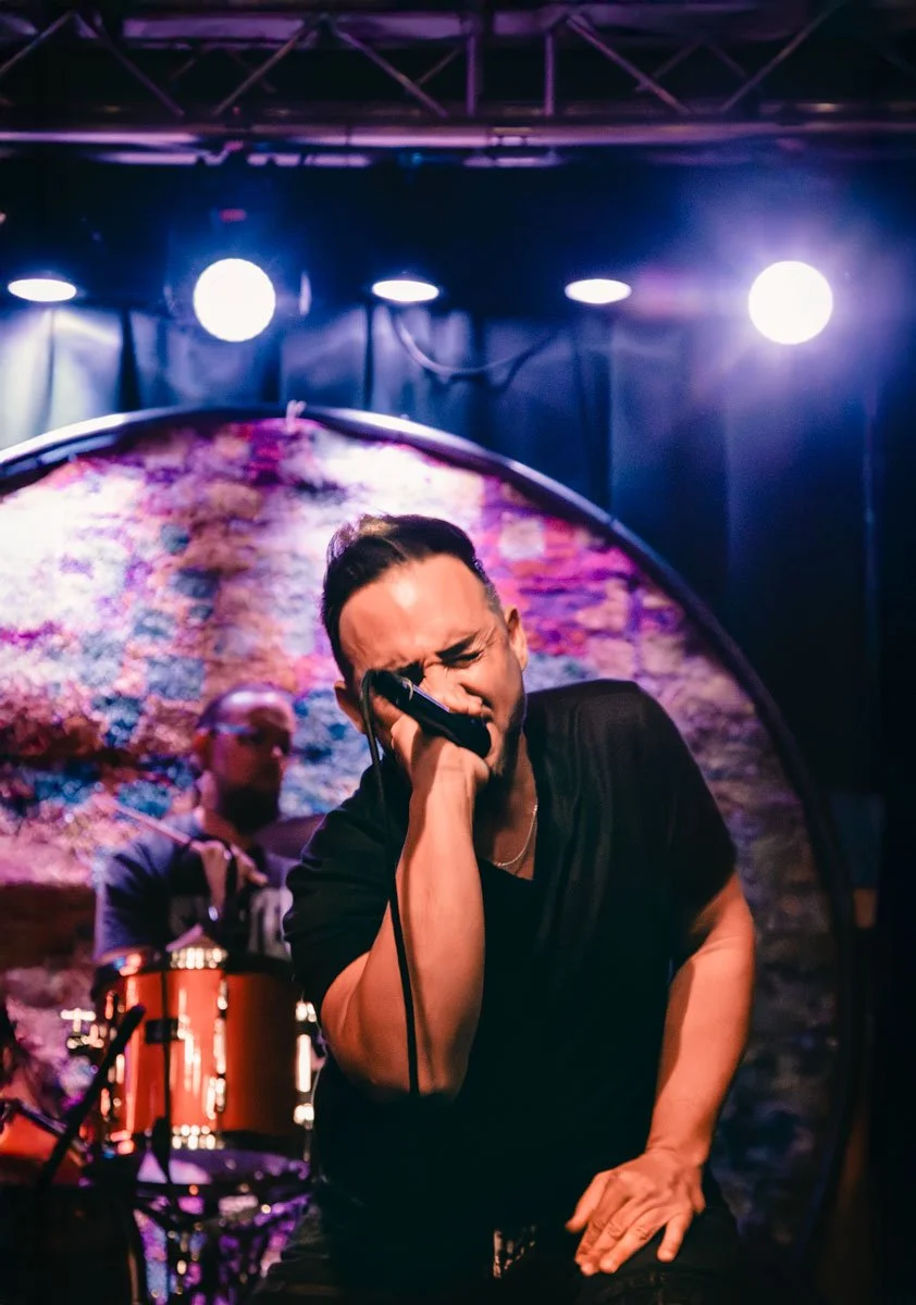 A man with dark hair and facial hair holding a microphone on stage, pointing towards the camera with stage lights and musical instruments in the background.