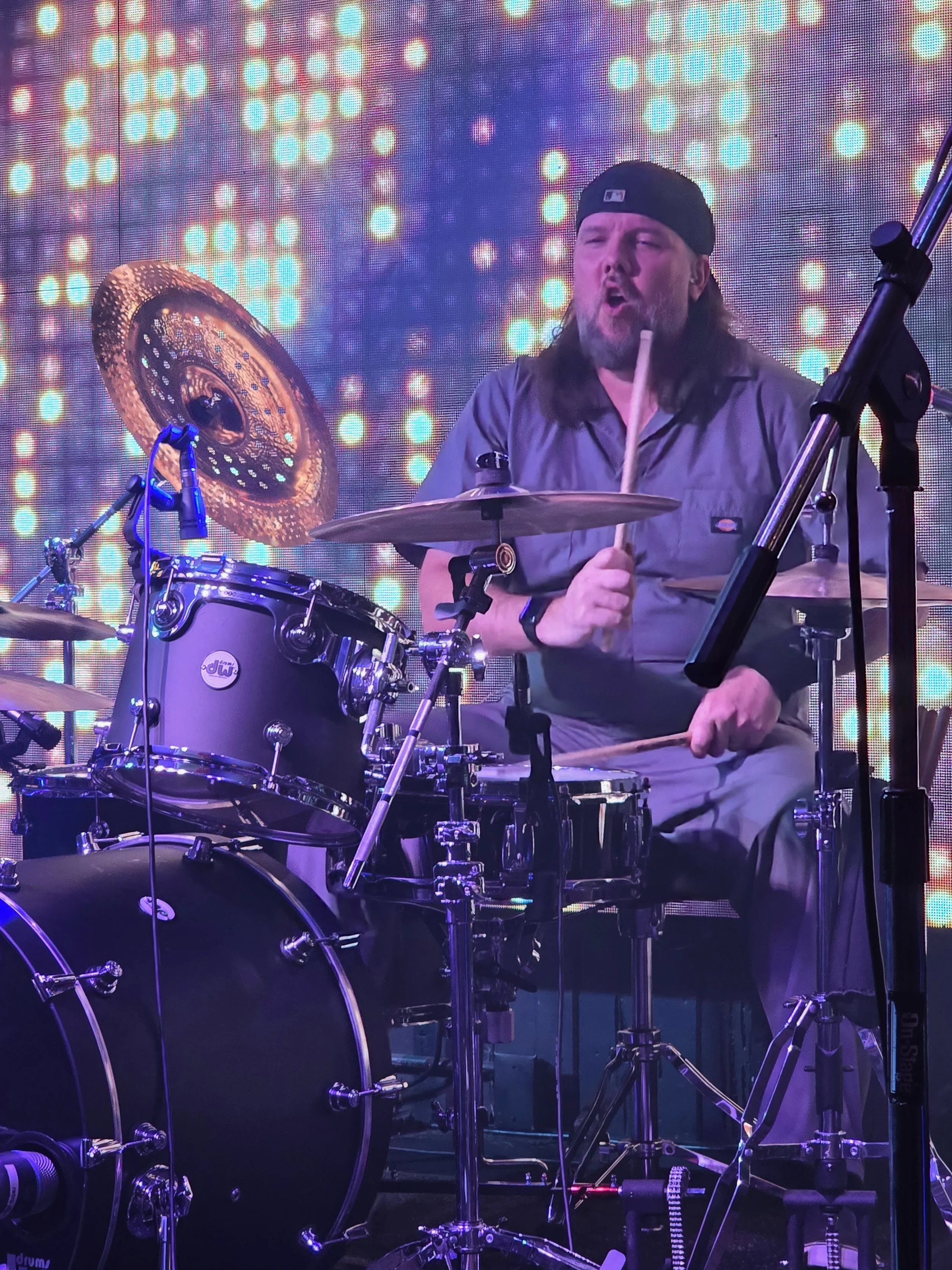 A drummer wearing a black cap and gray shirt playing a drum set with a colorful, illuminated background.