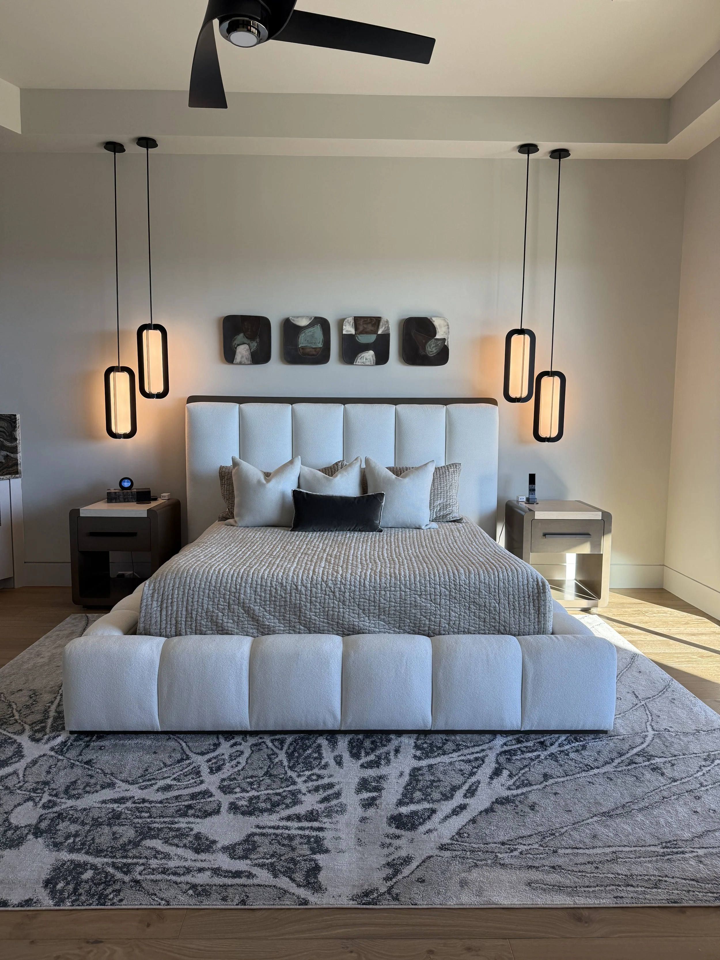 Modern bedroom with white upholstered bed, multiple pillows, black and beige quilt, abstract black and white wall art, contemporary hanging pendants above nightstands with decor, wooden floor, abstract blue/grey patterned rug, ceiling fan.