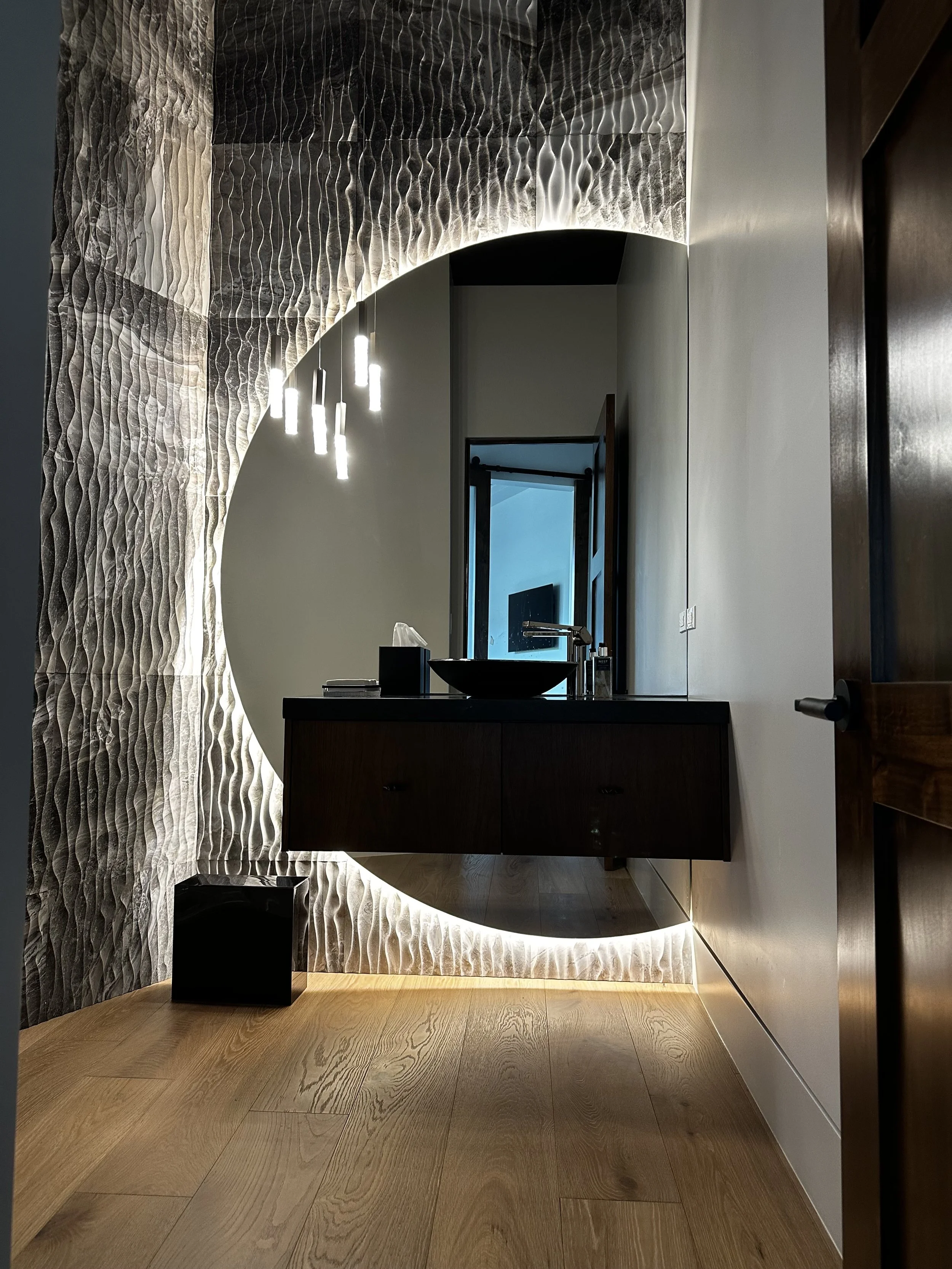 A warm modern bathroom with a floating round mirror, a floating dark walnut vanity with a black sink, and a textured wall with wave patterns. Pendant lights hanging from the ceiling and white oak flooring.