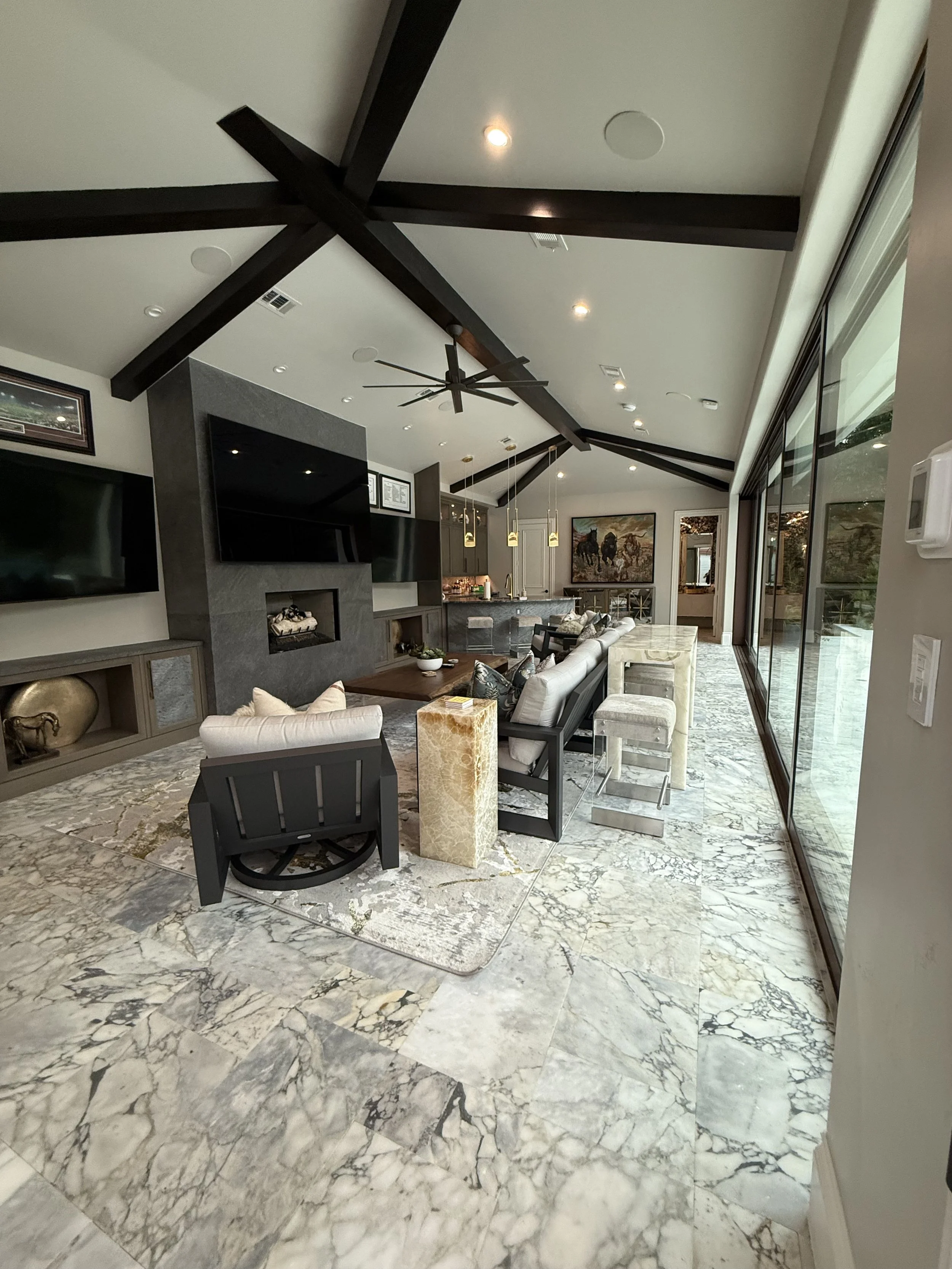 Spacious modern pool house/casita with a marble floor, large sliding glass pocket doors, a vaulted ceiling with dark knotty alder beams, a ceiling fan, a 3 large flat-screen TV 100", and a sectional sofa with decorative pillows.