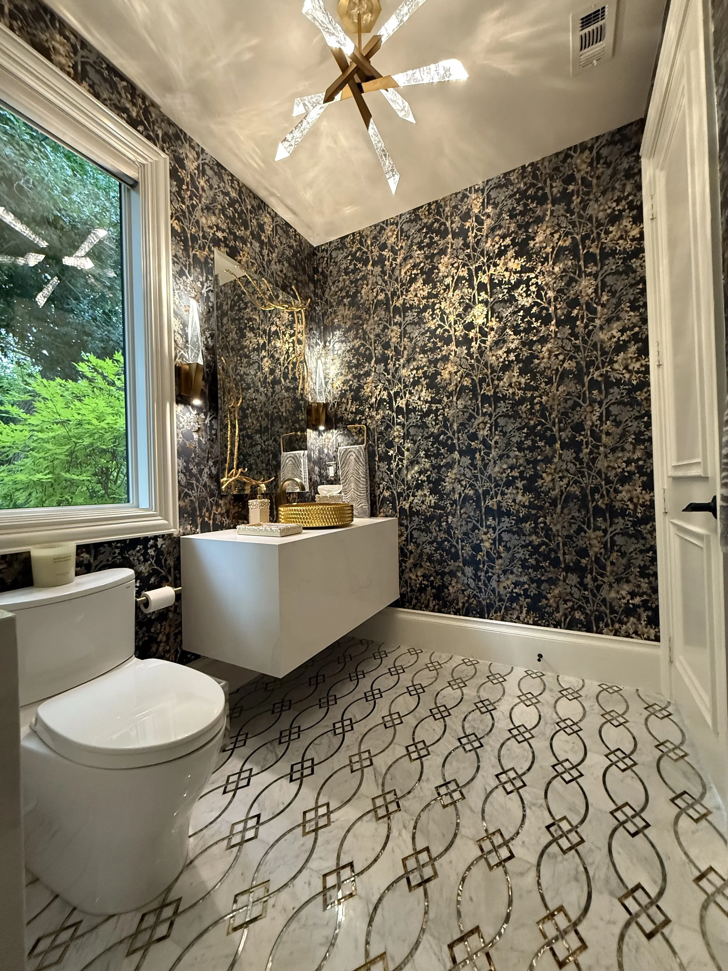 A modern bathroom with black and gold floral wallpaper, a large window with green trees outside, a white floating marble vanity, gold crystal chandelier, gold vessel sink and a mirror with gold branches coming out of it